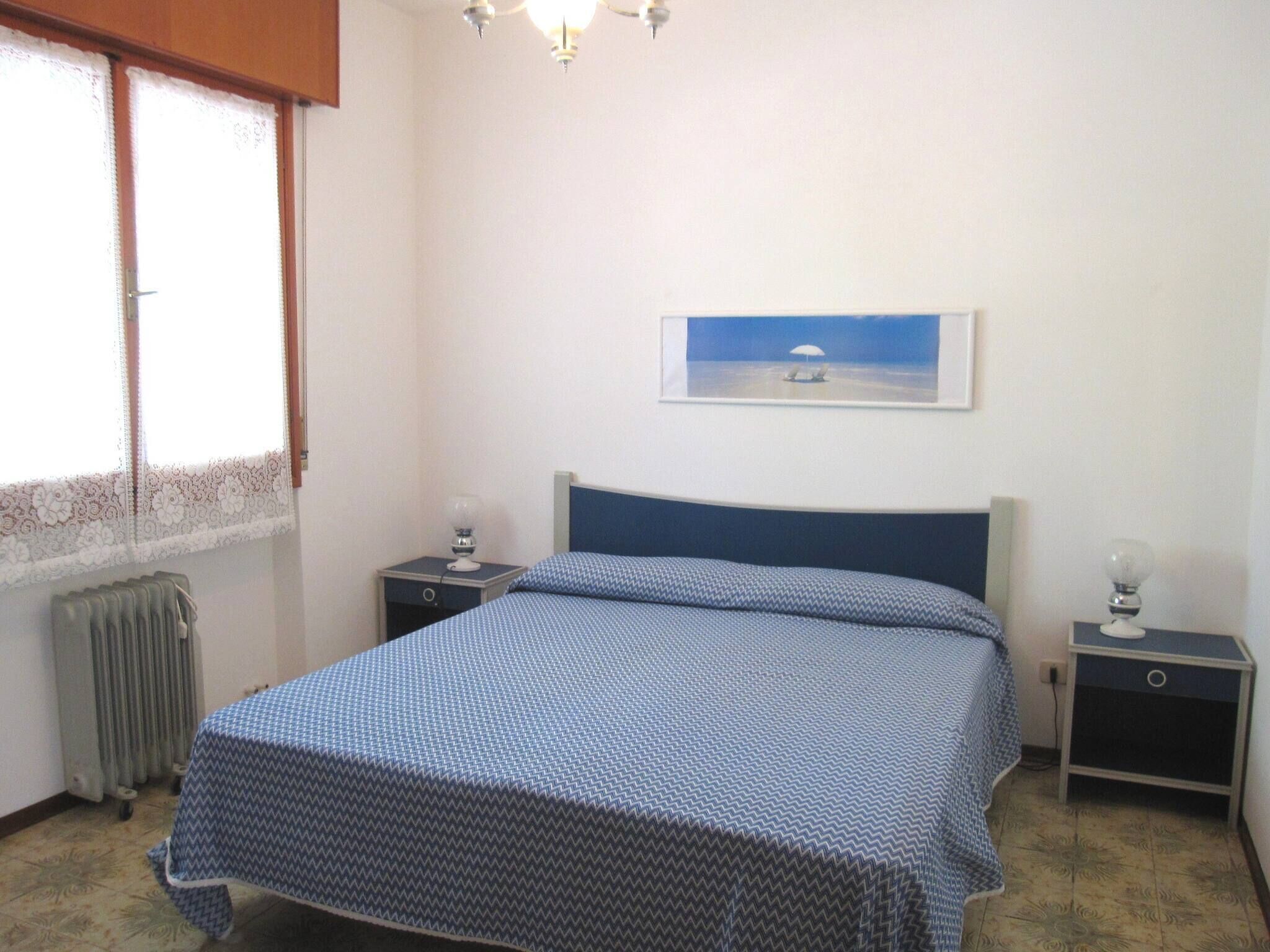 Un dormitorio con cama, dos mesitas de noche, un radiador, una ventana con cortinas de encaje y un cuadro en la pared.