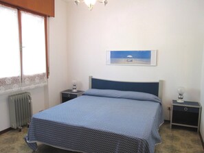 A bedroom with a bed, two bedside tables, a radiator, a window with lace curtains, and a framed picture on the wall. - Comfort With Terrace at Grado Pineta (Grado)
