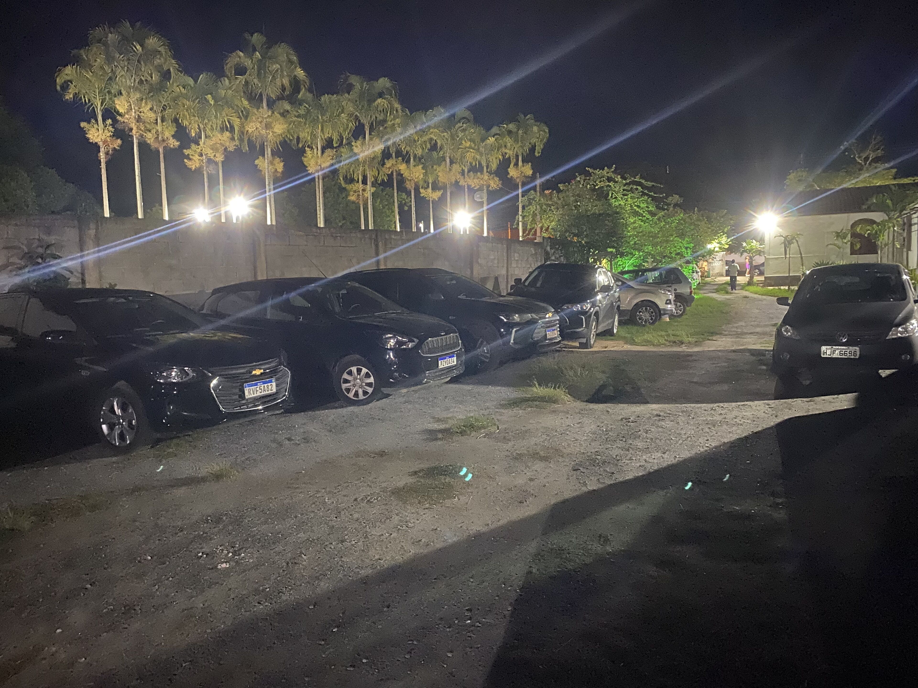 A nighttime parking area with several cars parked along a dirt path, surrounded by palm trees and illuminated by streetlights.