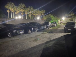A nighttime parking area with several cars parked along a dirt path, surrounded by palm trees and illuminated by streetlights.