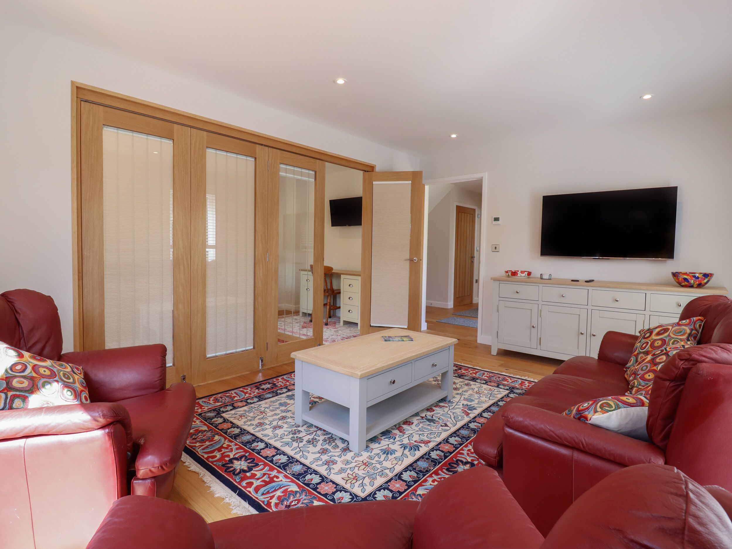Una sala de estar con sofás de cuero rojo, una mesa de centro blanca, un tapete estampado, un televisor montado en la pared y un mueble de madera.