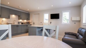 A modern kitchen with grey cabinets, a white countertop, and a built-in oven. There is a dining area with a round table and chairs, and a brown leather sofa. A flat-screen TV is mounted on the wall above the sofa. - Wellington Cottage (Ely)