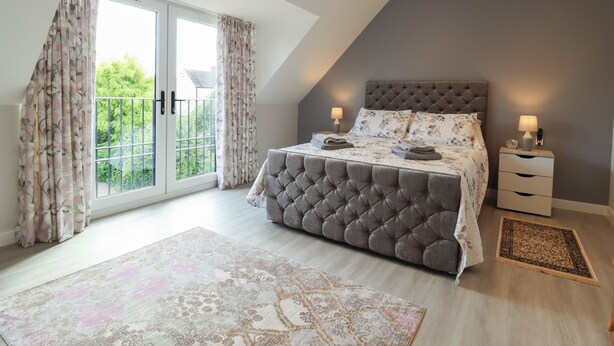 A bedroom with a large bed, two bedside tables, a window with curtains, and a view of greenery outside. - Wellington Cottage (Ely)