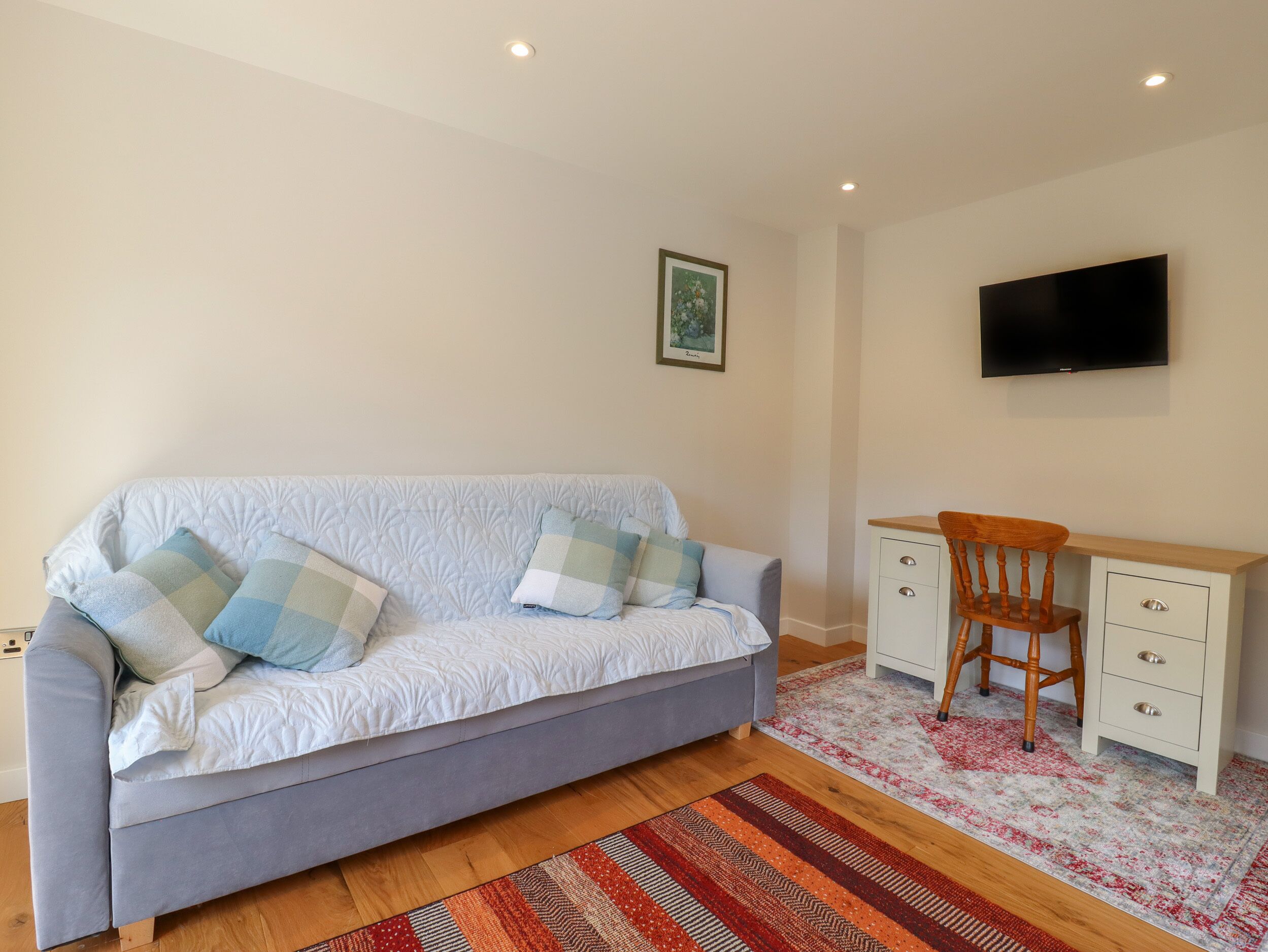 A room with a grey sofa, a wooden chair, a desk with drawers, a flat-screen TV mounted on the wall, and a patterned rug on the floor.
