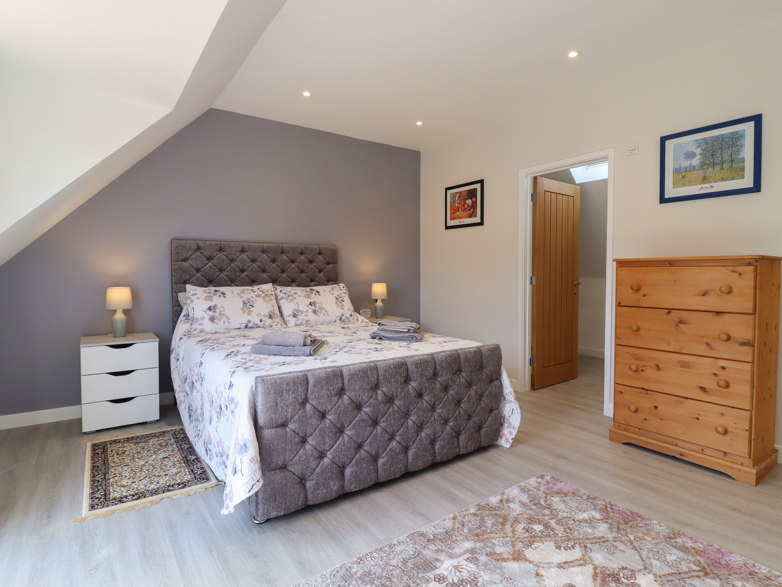 Un dormitorio con una cama, dos mesitas de noche, un aparador de madera y una puerta que da a otra habitación.
