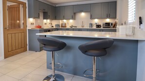 A modern kitchen with grey cabinets, a large island with two bar stools, and various appliances. - Wellington Cottage (Ely)