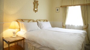Een tweepersoonsbed met witte lakens, een nachtkastje met een lamp, een spiegel met gouden lijst en een raam met gordijnen.