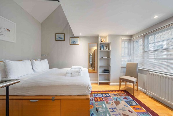 Une chambre à coucher avec un lit, une chaise, une bibliothèque et une fenêtre avec des stores.