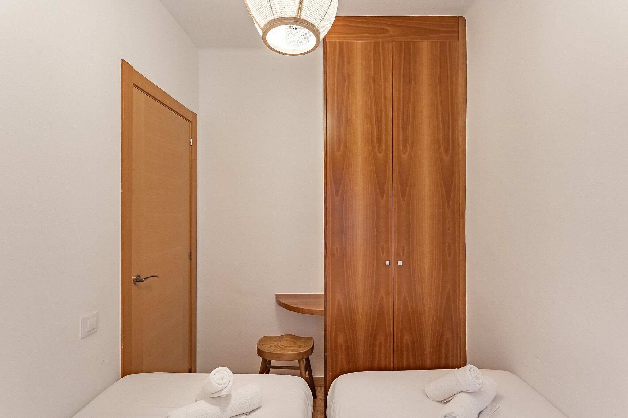 Quarto pequeno e bem arrumado, com duas camas, um guarda-roupa de madeira, um banquinho e uma prateleira na parede.