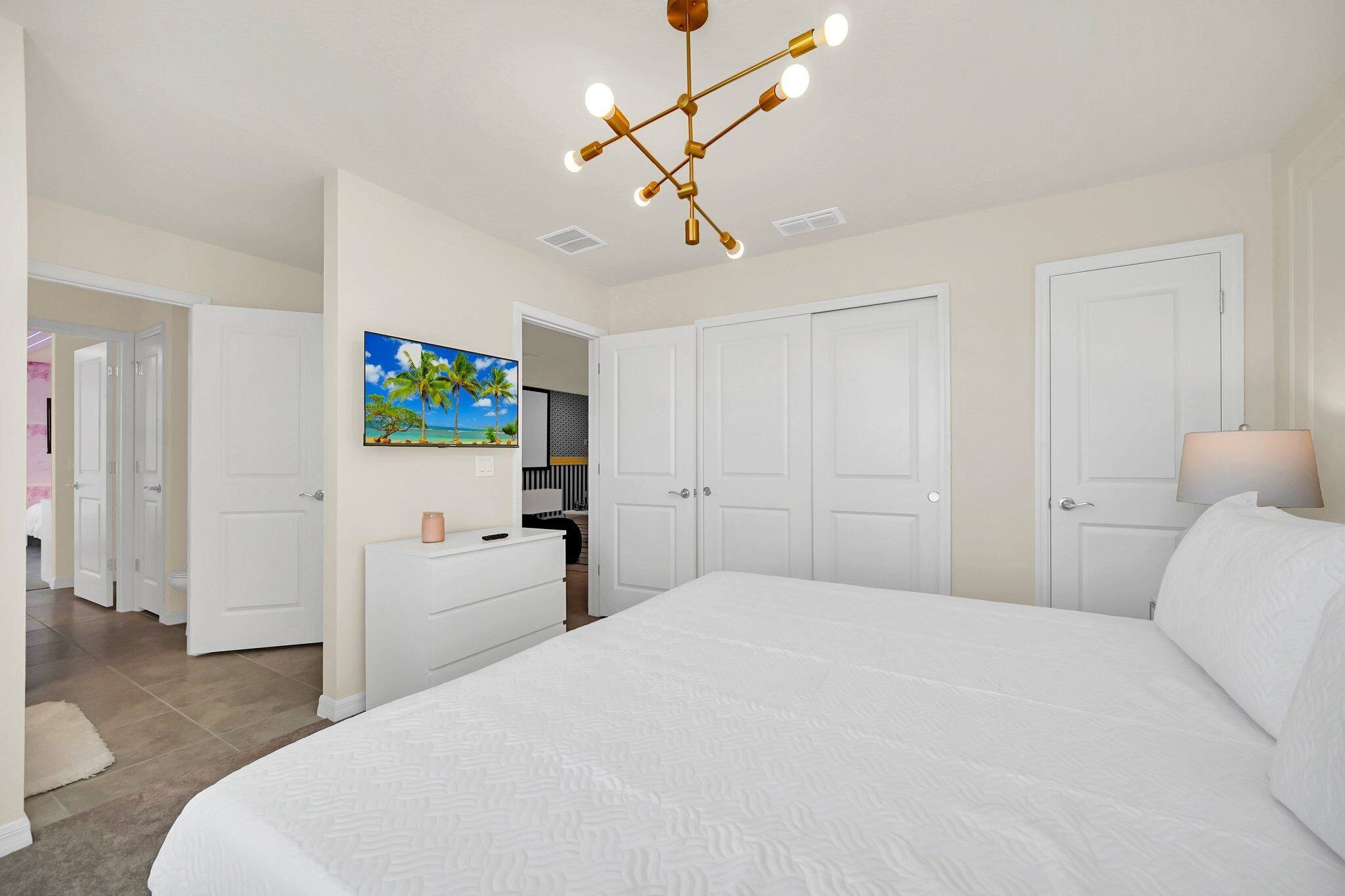 A bedroom with a large bed, white walls, a ceiling light fixture, and a painting of a beach scene.