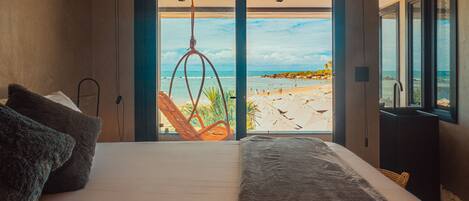Ein Schlafzimmer mit einem großen Bett, Blick auf einen Strand und einem Hängesessel.