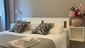Un lit bien fait, agrémenté d’oreillers à motifs, une table de chevet avec un vase de fleurs et un appareil de climatisation fixé au mur.
