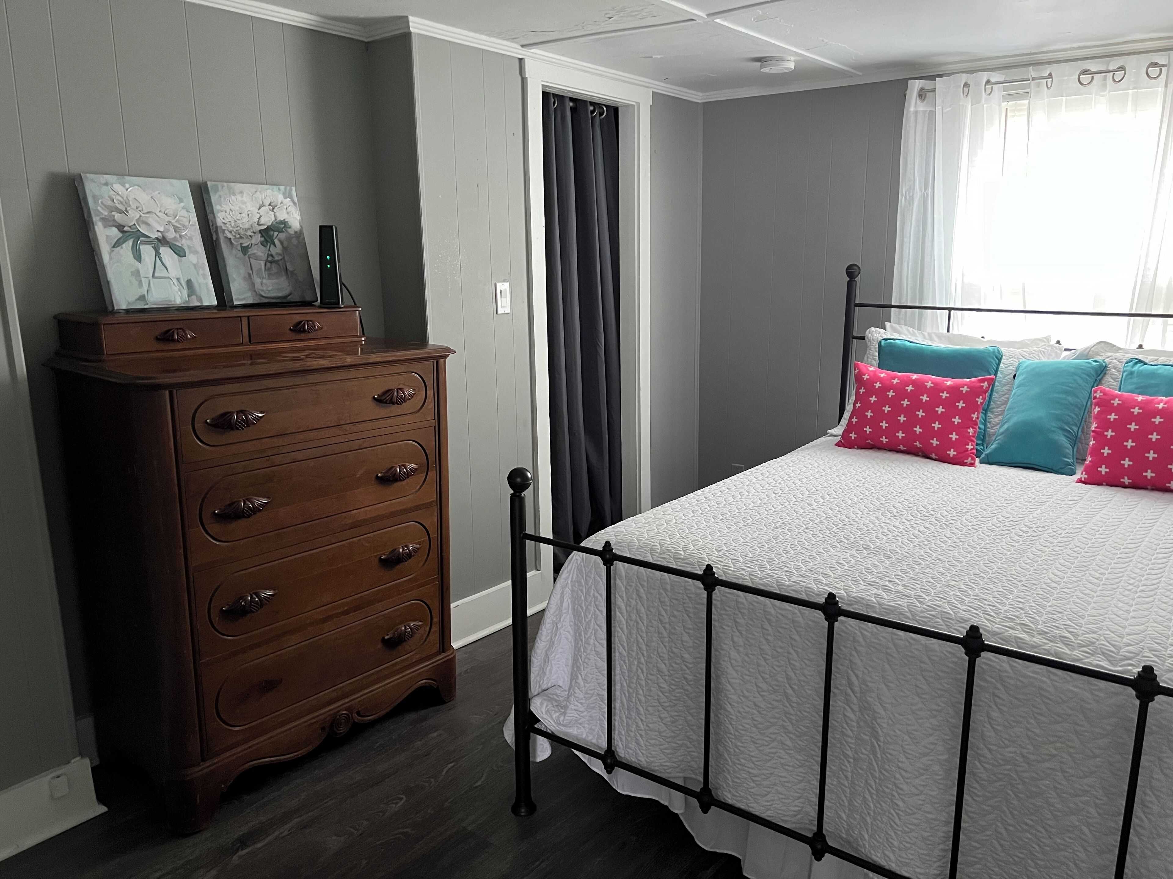 Un dormitorio con cómoda de madera, cama con ropa de cama blanca y almohadas estampadas, ventana con cortinas y puerta con cortinaje.
