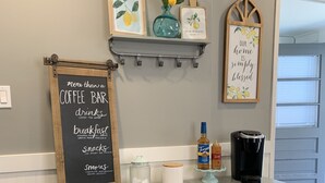 Un coin cuisine avec un tableau noir indiquant les boissons chaudes (café, thé), l’eau, le petit-déjeuner, les en-cas et les « s’mores ». On y trouve des paniers étiquetés « en-cas » et « petit-déjeuner » sur des étagères, une machine à café et une bouteille de sirop Torani.