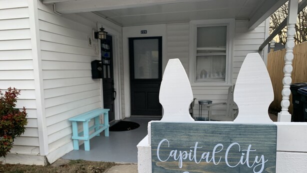 Une pancarte en bois portant l’inscription « Capital City Cottage » est placée devant une clôture blanche à piquets.