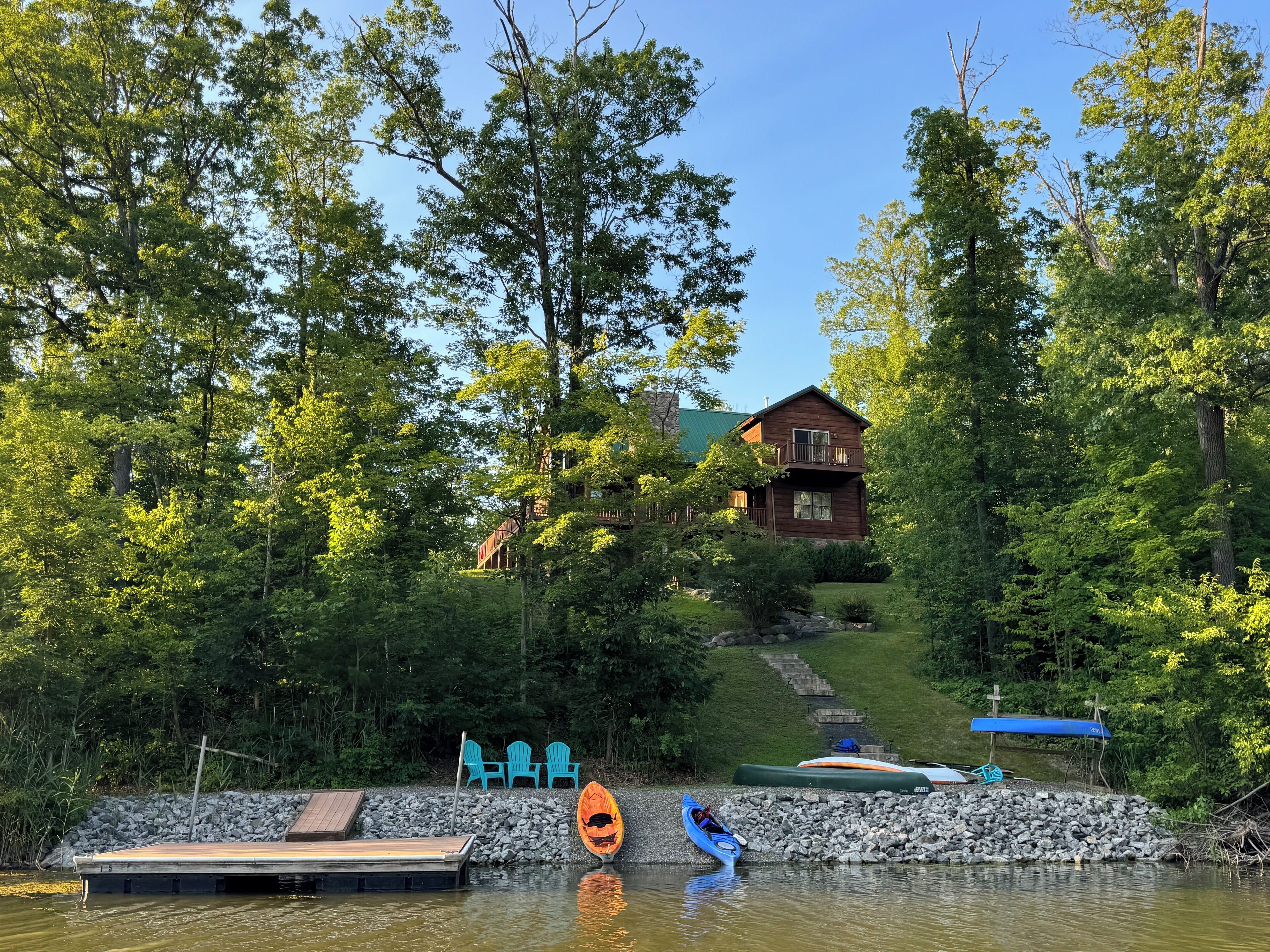 Une cabane entourée d’une forêt dense, une jetée avec des chaises et des kayaks.