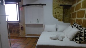 A bedroom with a bed, white pillows, a black and white patterned pillow, a towel, a shelf with a bottle, and a fire extinguisher. - Authentic Escape: Tosca Stone Studio in Javea (Xàbia)