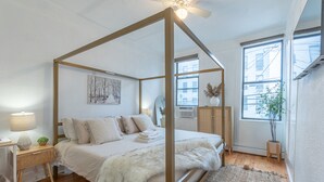 A bedroom with a four-poster bed, a bedside table with a lamp, a small plant, and a window with a view of buildings. - Spring 25% Discount is here, 1King bed, Near NYC (Hoboken)