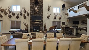 A spacious living room with a stone fireplace, mounted animal heads, and a dining area with a large wooden table and upholstered chairs. - Spacious modern lodge outside of city (Grand Island)