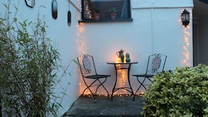 Un pequeño espacio al aire libre con dos sillas de metal y una mesa, rodeado de plantas y luces colgantes.