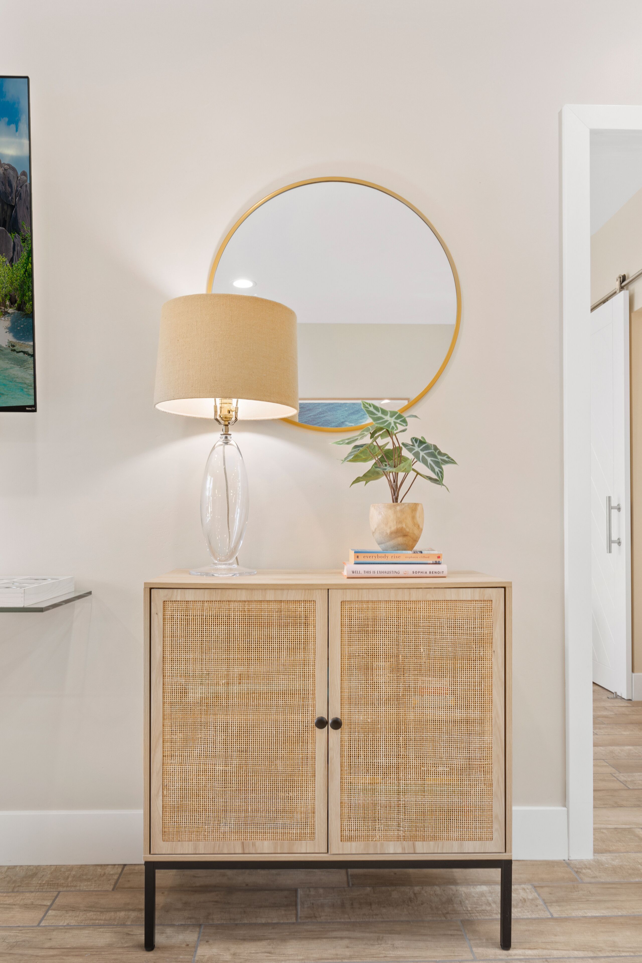 Ein Holzschrank mit Rattanfront, eine klare Glaslampe mit beiger Lampenschirm, ein runder Spiegel und ein kleiner Blumentopf mit Büchern darauf.