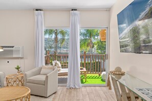 A modern living room with a sofa, a glass table, and a view of a green outdoor area with a wooden deck and palm trees.