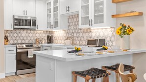 Une cuisine moderne avec des meubles blancs, un îlot central, des tabourets en bois, un éclairage suspendu et un revêtement mural décoratif à motifs géométriques.