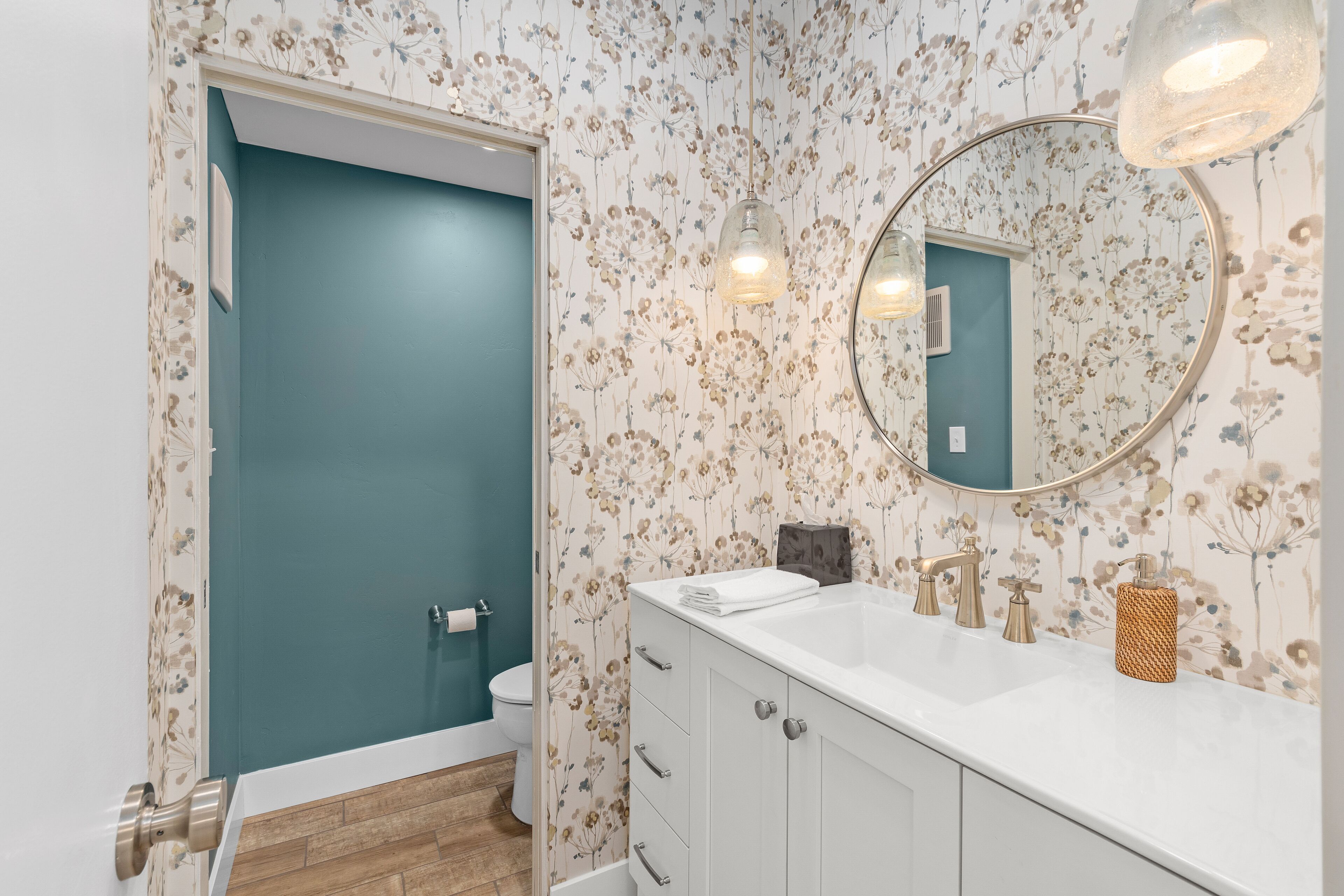 Un baño con pared color turquesa, papel tapiz floral, un lavamanos blanco con un espejo redondo y suelo de madera.