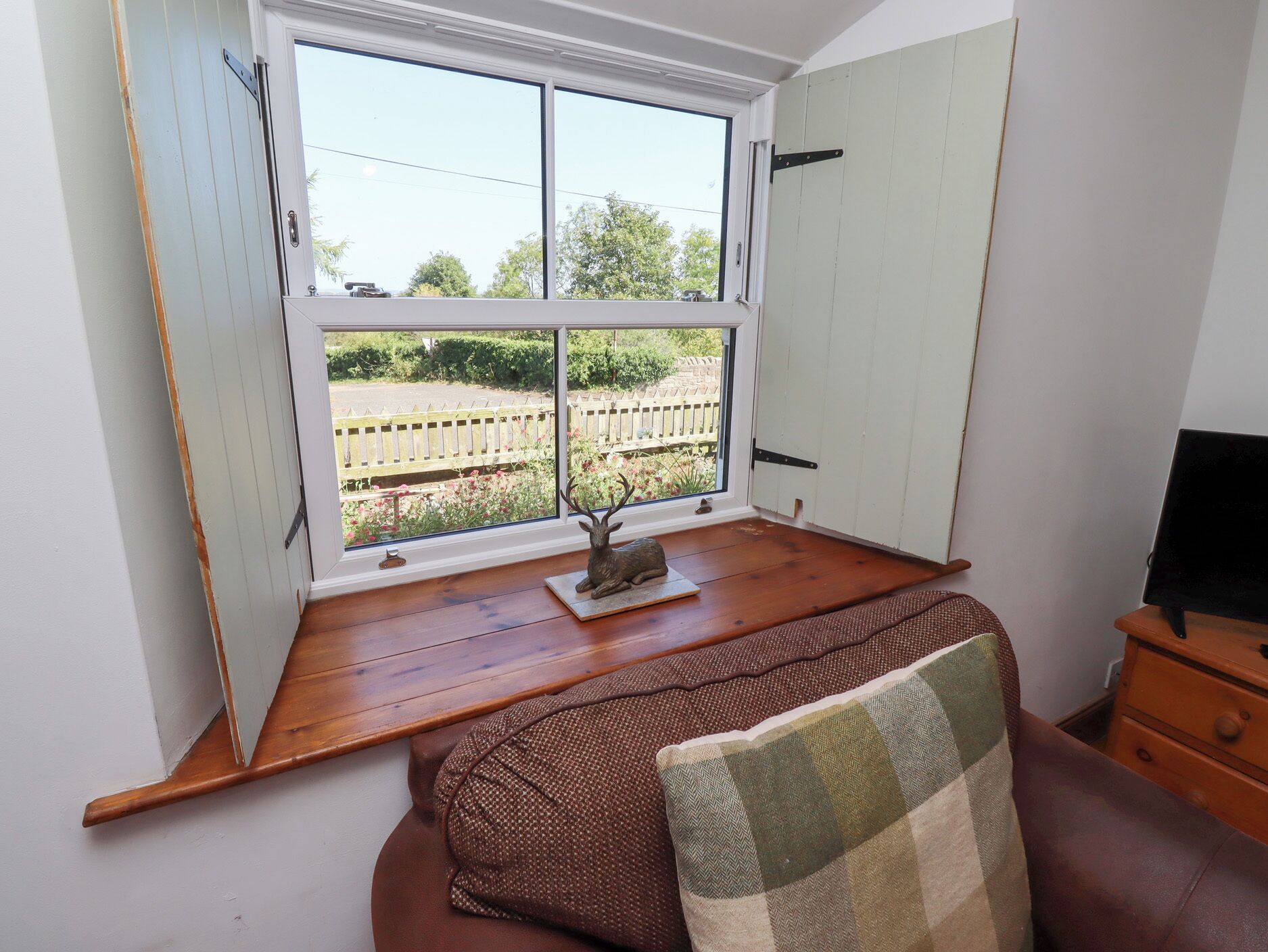 A room with a wooden floor, a window with a view of a garden, a small table with a decorative item, and a brown sofa with a patterned throw.