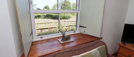 A room with a wooden floor, a window with a view of a garden, a small table with a decorative item, and a brown sofa with a patterned throw.