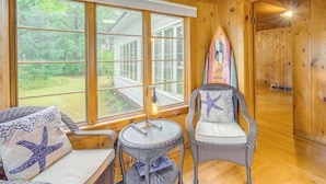 Une pièce chaleureuse, meublée de mobilier en osier, avec une planche de surf et une grande fenêtre donnant sur un jardin.