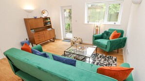 A modern living room with a green sofa, a teal armchair, and a glass coffee table. There is a wooden cabinet with books and decorative items, and a window with a view of greenery outside. - James' Place (Falmouth)