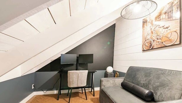 Ein modernes Schlafzimmer im Loftsstil mit schrägem Dach, einer grauen Couch, einem kleinen Schreibtisch mit Stuhl, einem Flachbildfernseher und einer Wandleuchte.
