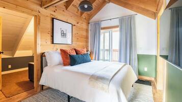 Une chambre confortable avec un plafond en bois, un lit recouvert de draps blancs et agrémenté de coussins colorés, un tableau encadré au mur et une grande fenêtre avec des rideaux.