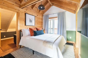 Ein gemütliches Schlafzimmer mit Holzbalkendecke, einem Bett mit weißer Bettwäsche und bunten Kissen, einem gerahmten Bild an der Wand und einem großen Fenster mit Vorhängen.