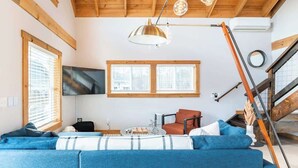 Ein modernes Wohnzimmer mit einem blauen Modulsessel, einem Couchtisch und einem Fernseher. Große Fenster, eine Treppe und Holzbalken zieren die Decke.