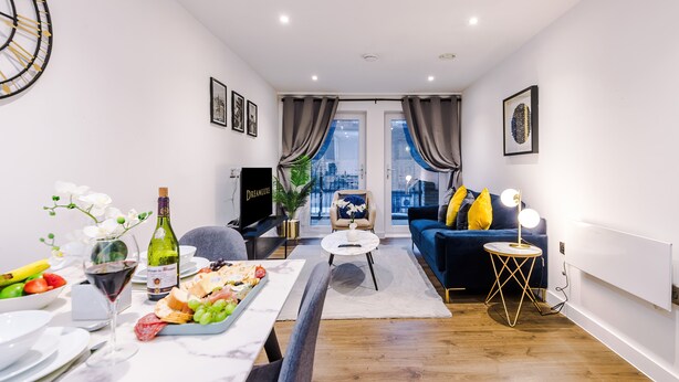 A modern living room with a dining area, featuring a sofa, a TV, a dining table with food, and a view of the city through large windows. - Impeccable 2-bed Apartment in Salford (Salford)