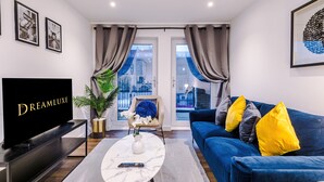 A modern living room with a large flat-screen TV, a blue sofa with yellow and gray cushions, a white coffee table with a vase and remote, and a city view through large windows. - Impeccable 2-bed Apartment in Salford (Salford)