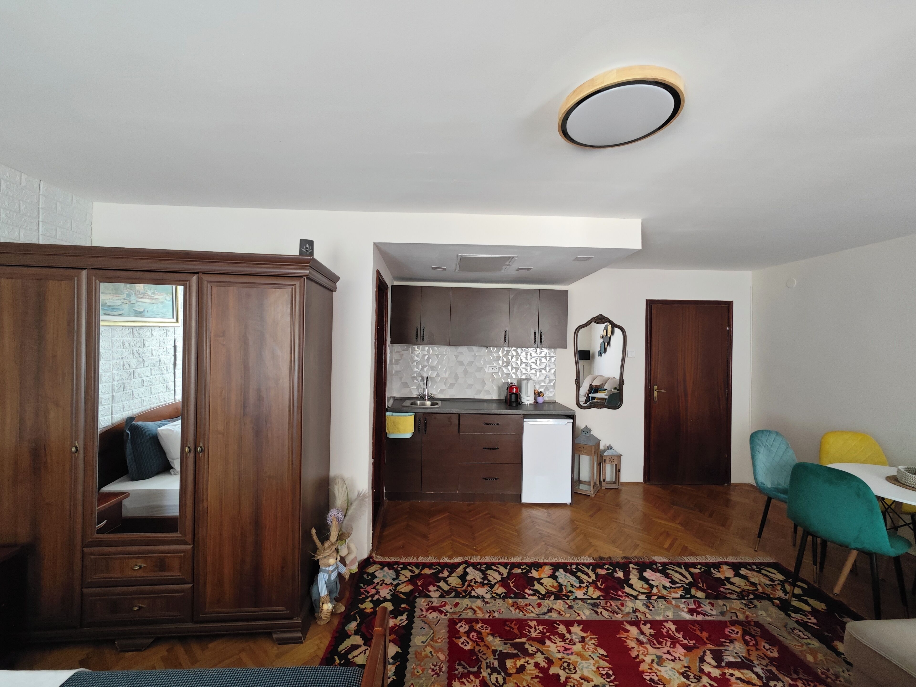 A room with a wooden wardrobe, a bed, a small rug, and a view into a kitchen area with cabinets and a sink.