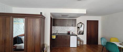 A room with a wooden wardrobe, a bed, a small rug, and a view into a kitchen area with cabinets and a sink.