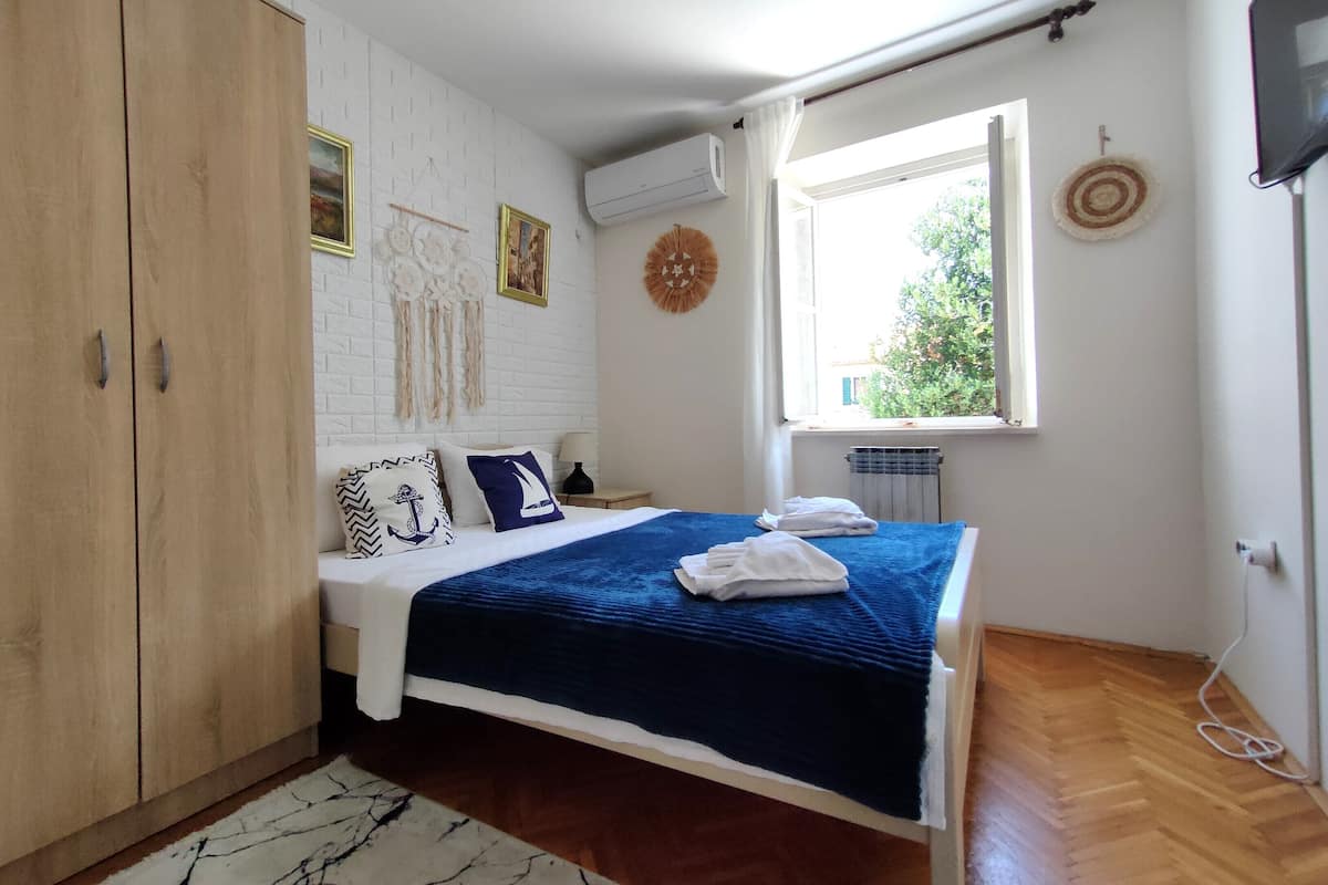 A bedroom with a bed, wooden wardrobe, white brick wall, air conditioning unit, window with view, and a wall decoration.