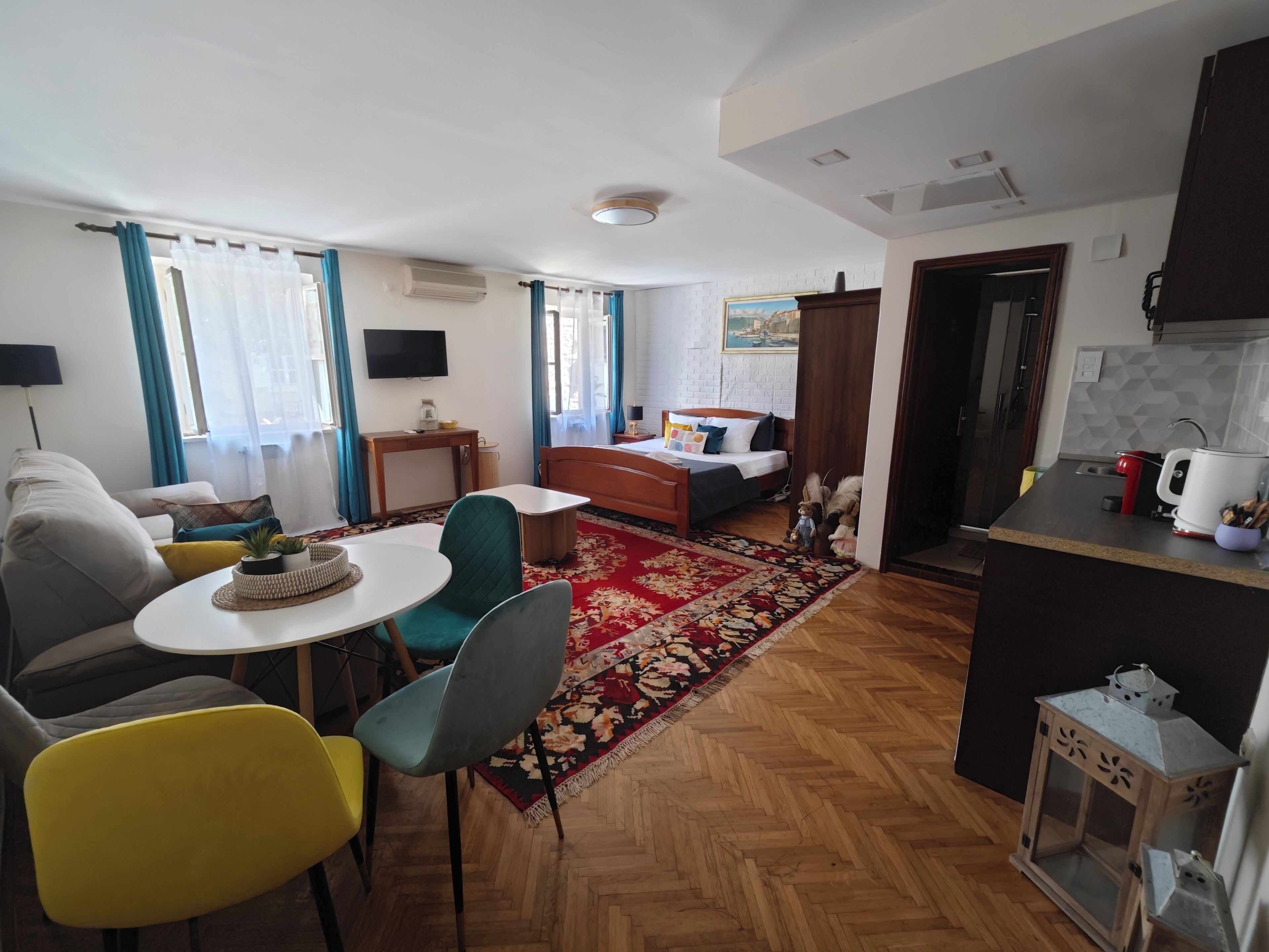 A modern hotel room with a bed, a dining area, a kitchen, and a TV.