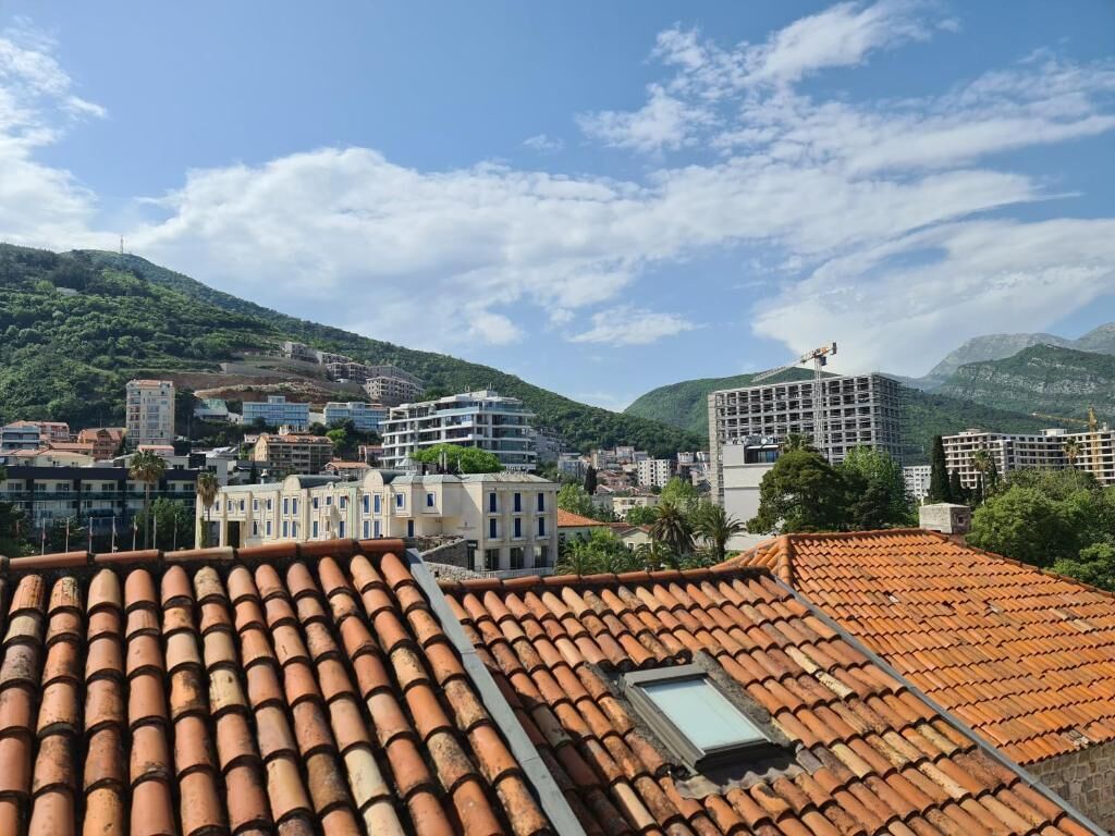 A cityscape with terracotta-tiled rooftops, a mix of modern and traditional buildings, and a mountainous backdrop.