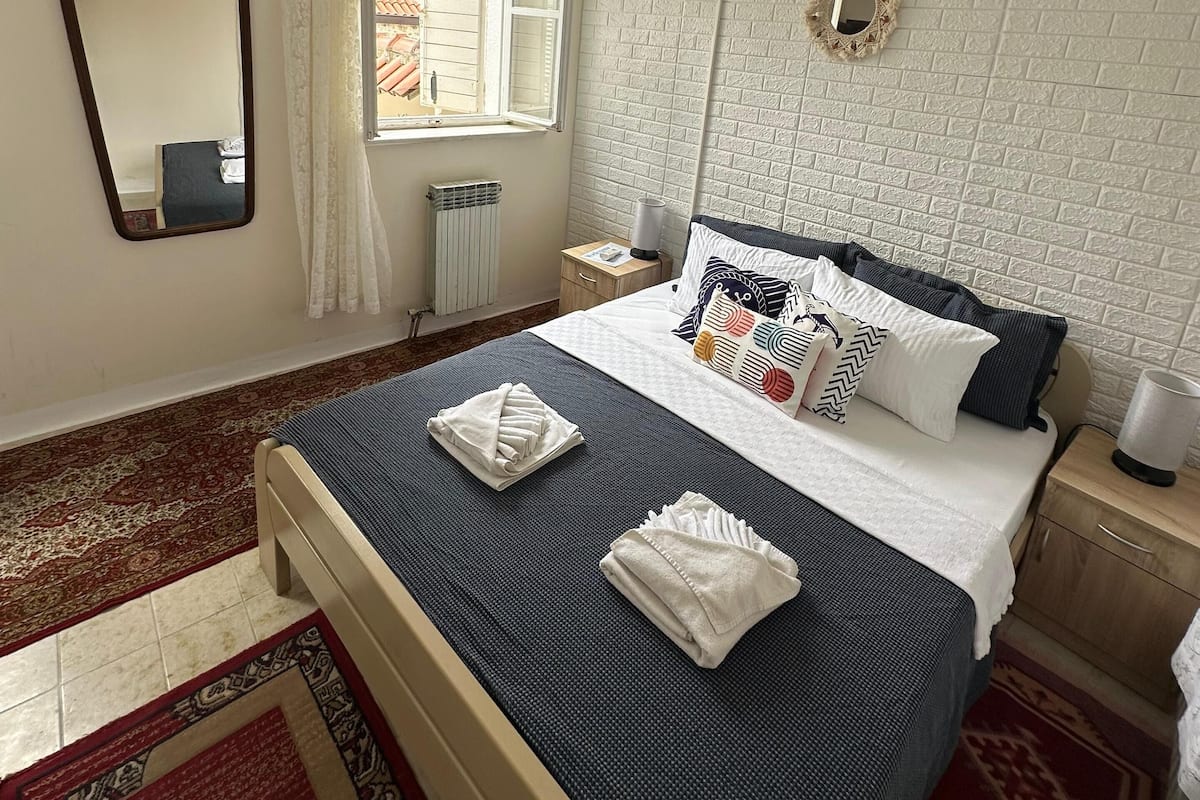 A neatly arranged bedroom with a bed, a bedside table, a mirror, and a window with curtains.