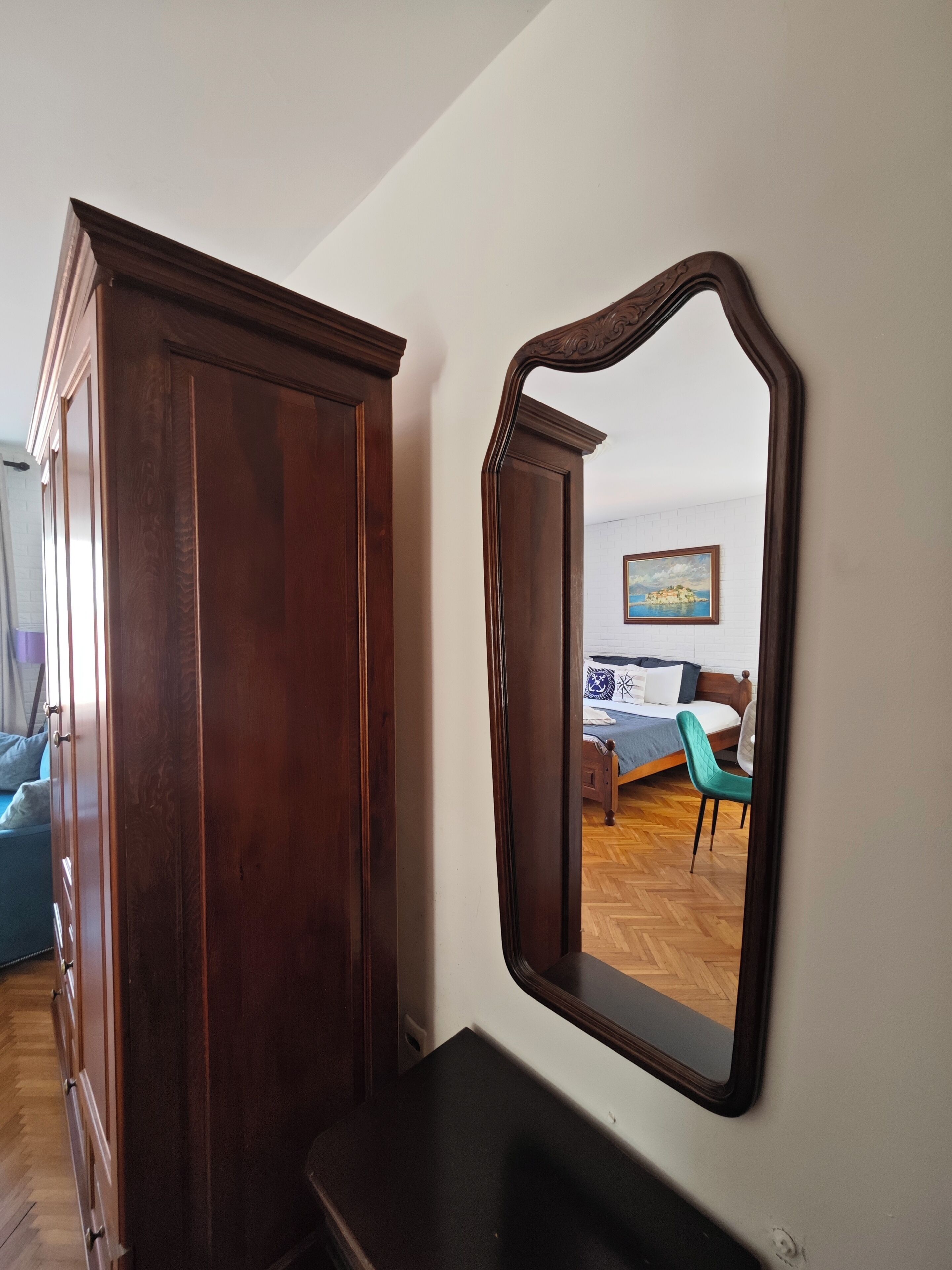 A room with a wooden wardrobe, a mirror, a bed with a blue bedspread, a green chair, and a wooden floor.