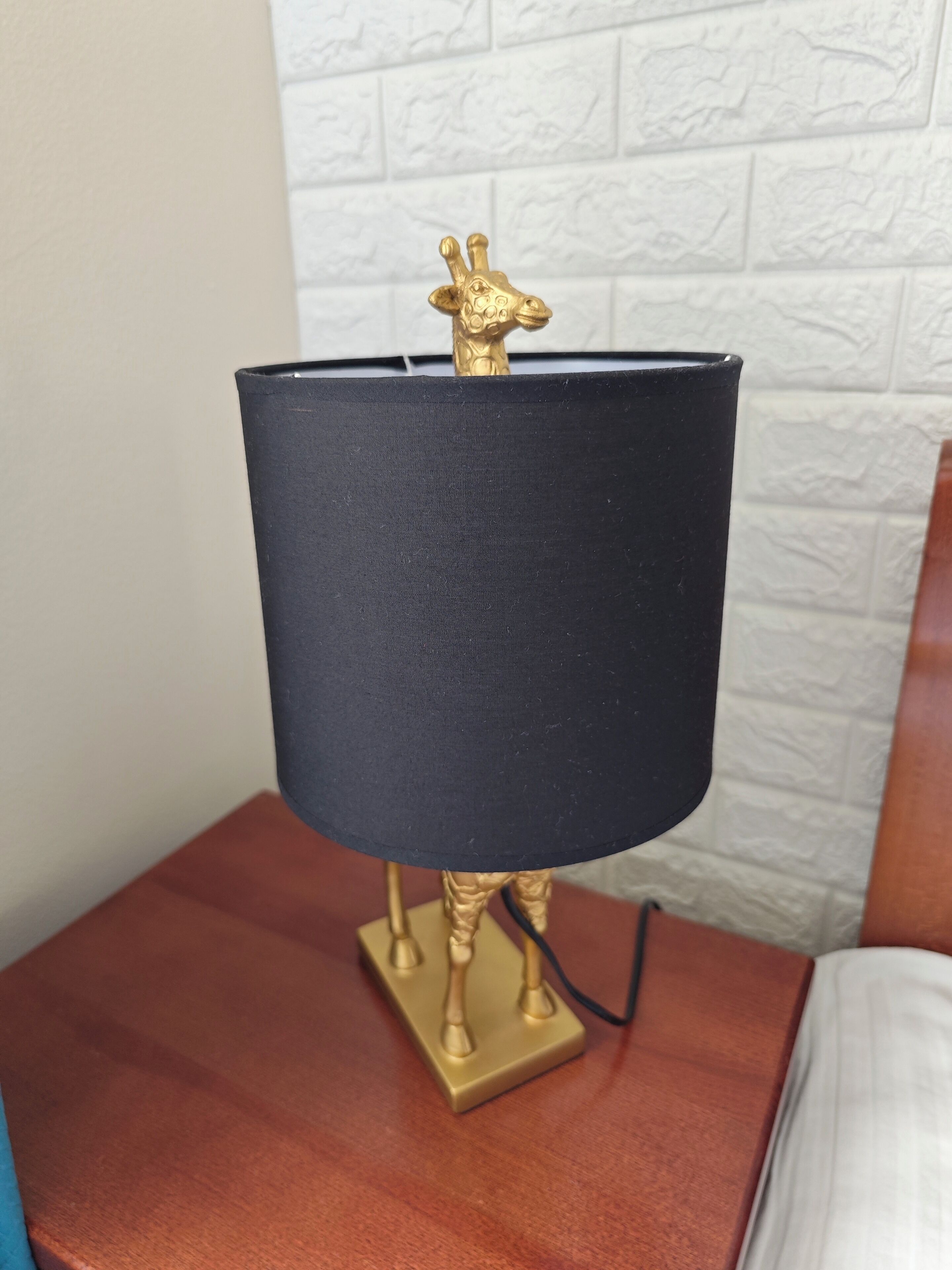 A table lamp with a black shade and a golden giraffe figurine on top, placed on a wooden nightstand.