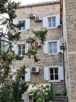Ein Gebäude aus Stein mit zahlreichen Fenstern, von denen einige mit Fensterläden versehen sind, und mehreren Klimaanlagen an der Außenwand.