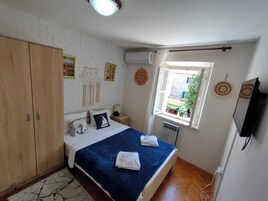 Un dormitorio con cama, armario de madera, pared de ladrillo blanco, unidad de aire acondicionado, ventana con cortinas y televisor de pantalla plana.