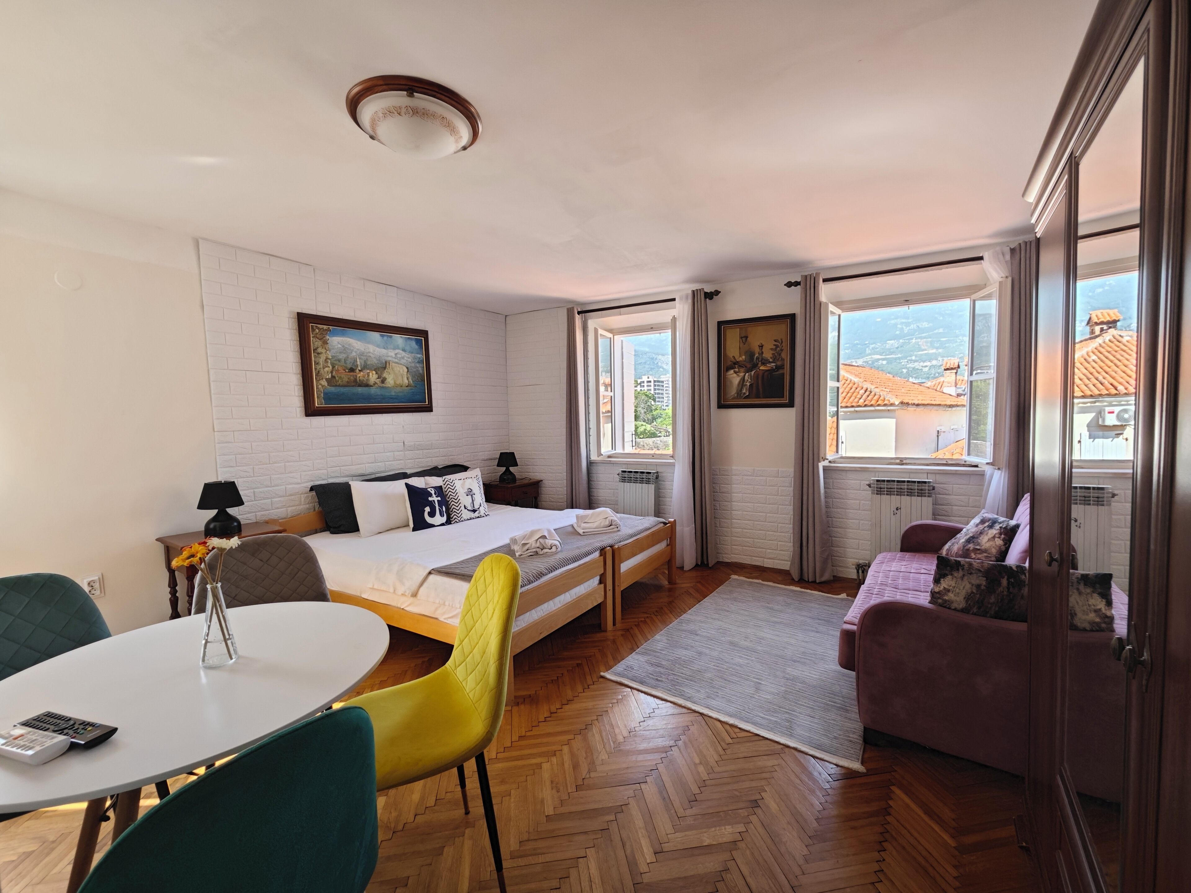A bedroom with a bed, a desk with a chair, a sofa, and a window with a view of buildings.