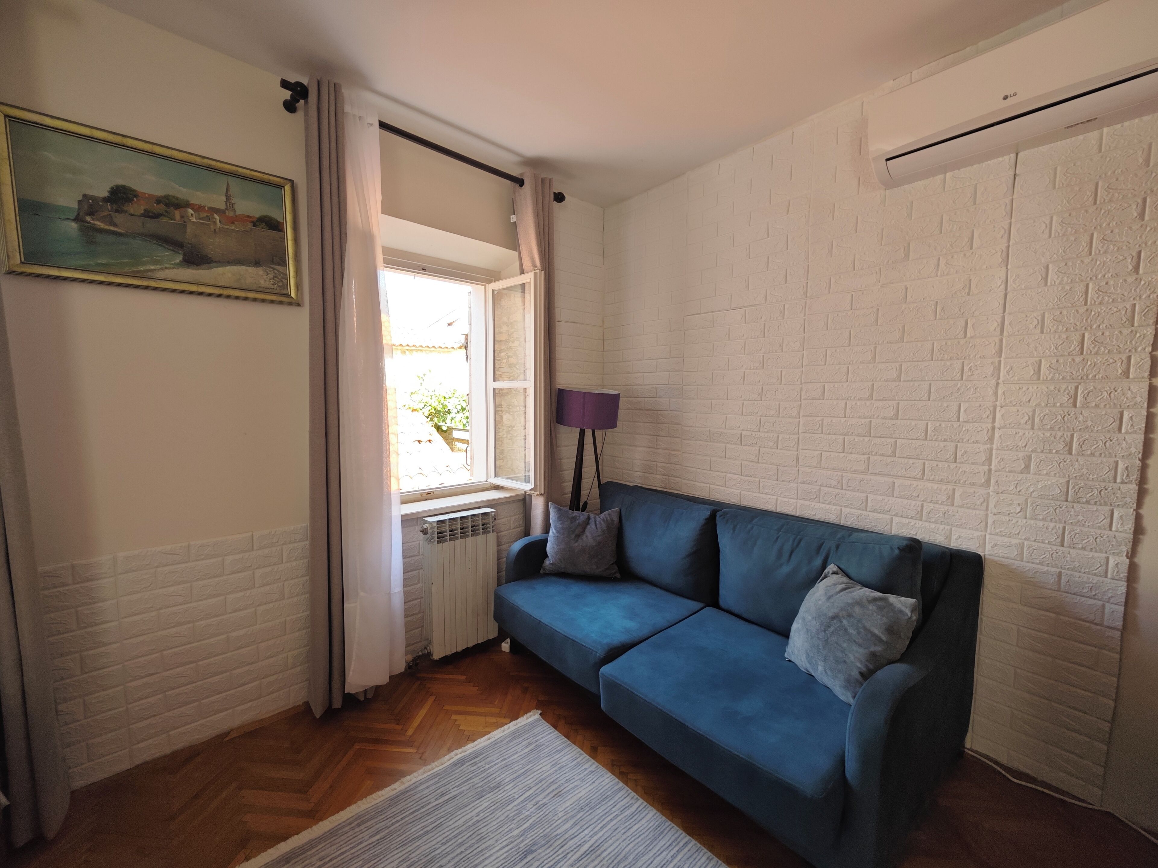 Una habitación con un sofá azul, un radiador, una pared de ladrillo blanco, una ventana con cortinas y un cuadro en la pared.